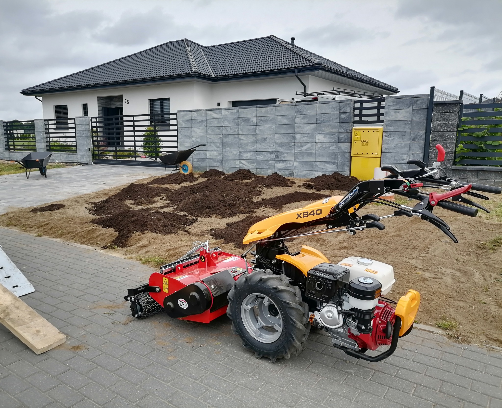 Żółto-czerwony glebogryzarek z silnikiem spalinowym na kostce brukowej, przygotowujący teren pod trawnik przed nowoczesnym domem z szarą kostką i metalowym ogrodzeniem.