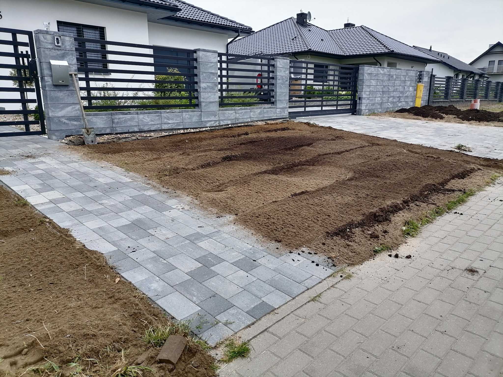 Przygotowany teren pod ogród przed nowoczesnym domem z szarą kostką brukową i czarnym ogrodzeniem, widoczna łopata oparta o słupek ogrodzenia.