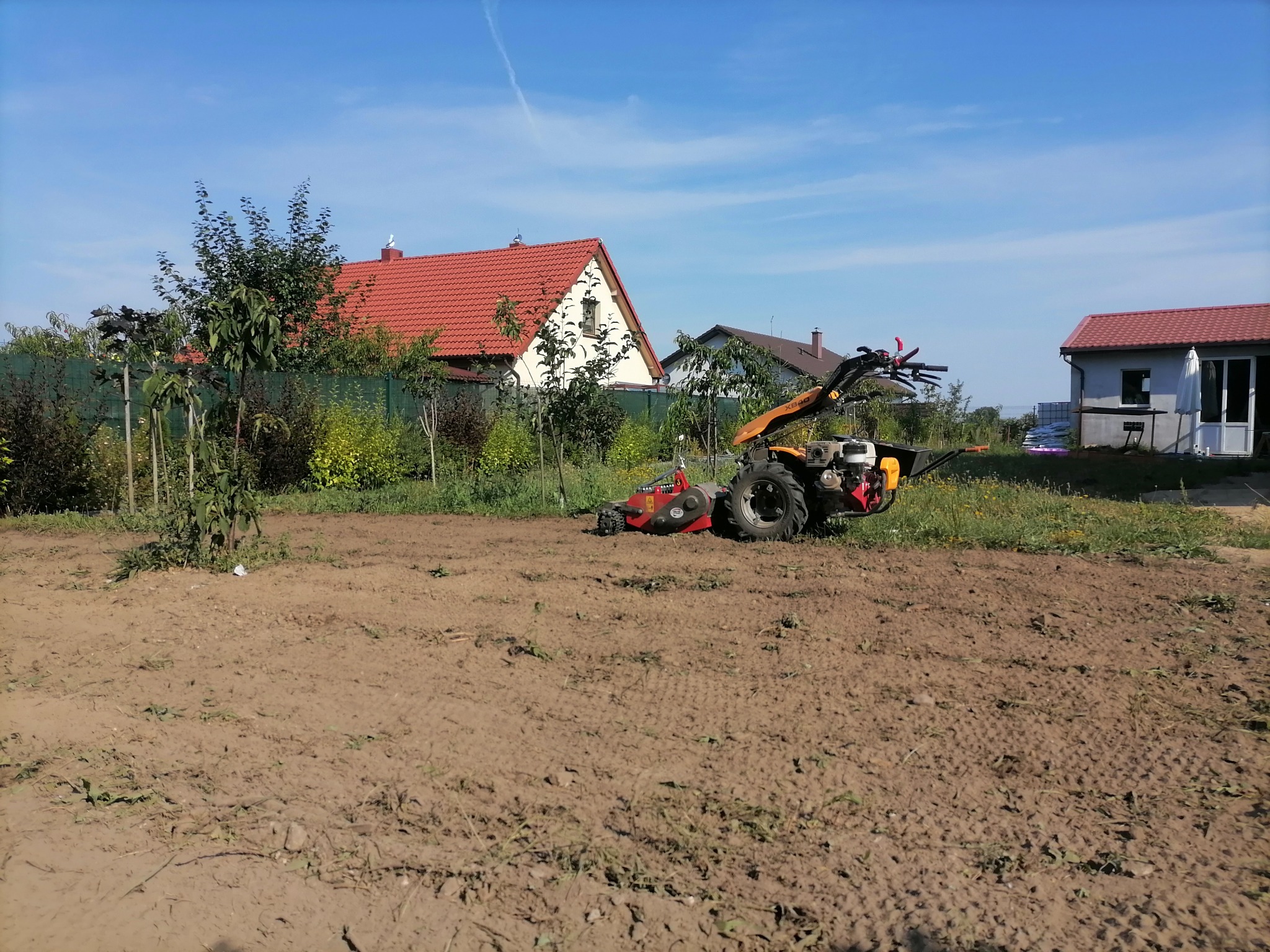 Glebogryzarka spalinowa w trakcie pracy na polu, przygotowująca ziemię pod uprawę. W tle widoczne budynki mieszkalne i drzewa owocowe.