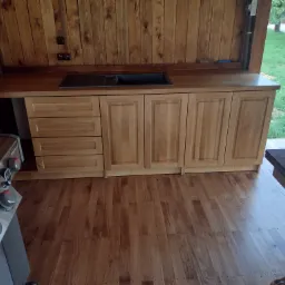 Drewniana zabudowa kuchenna z blatem i zlewem, widoczna część grilla, drewniana podłoga i ściany w zewnętrznej kuchni.