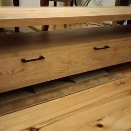 Stolik RTV z drewna dębowego z trzema szufladami, na czarnych metalowych nogach, na podłodze z desek.