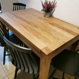 Stół z jasnego drewna z grubym blatem i nogami, otoczony czarnymi krzesłami, na blacie dekoracja z wrzosu w metalowej osłonce.