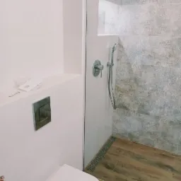 Nowoczesna łazienka z kabiną prysznicową typu walk-in, wykończona płytkami imitującymi kamień i drewnopodobną podłogą, widoczny biały sedes podtynkowy i bateria prysznicowa.