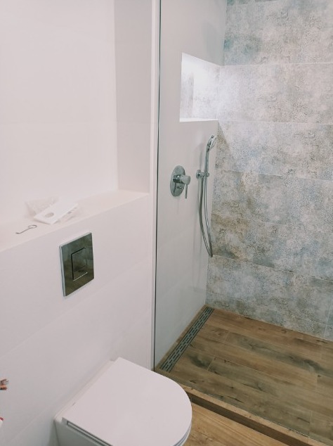 Nowoczesna łazienka z kabiną prysznicową typu walk-in, wykończona płytkami imitującymi kamień i drewnopodobną podłogą, widoczny biały sedes podtynkowy i bateria prysznicowa.