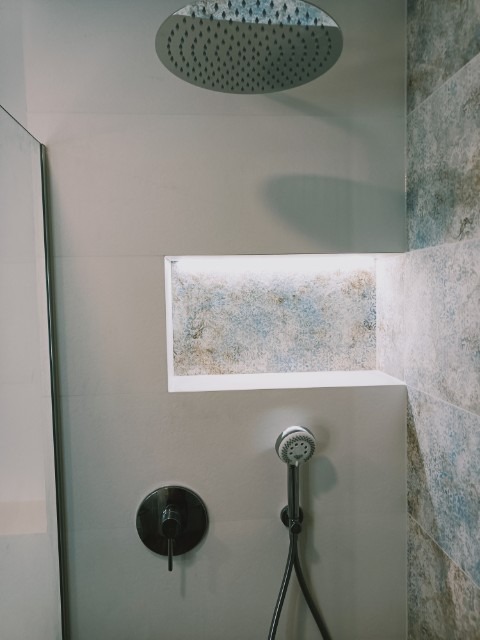 Nowoczesny prysznic z deszczownicą, wnęką z podświetleniem i baterią w kolorze czarnym na tle płytek imitujących marmur.