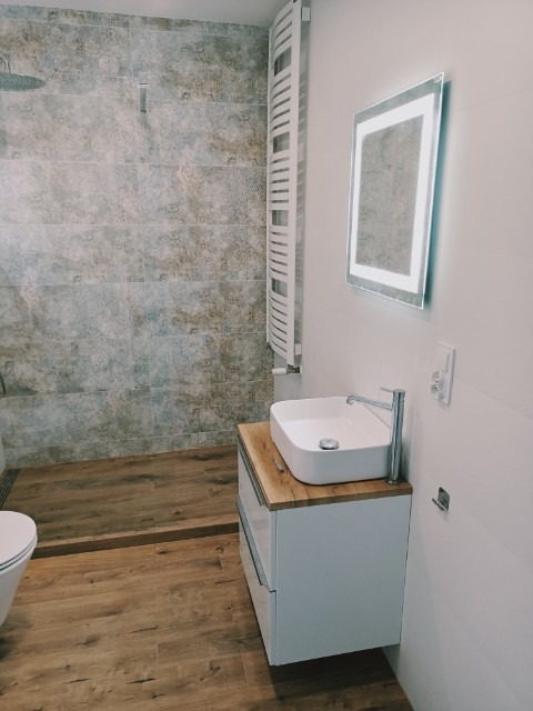 Nowoczesna łazienka z drewnopodobnymi płytkami podłogowymi i ściennymi, białą umywalką nablatową na szafce z drewnianym blatem, podświetlanym lustrem i grzejnikiem drabinkowym.