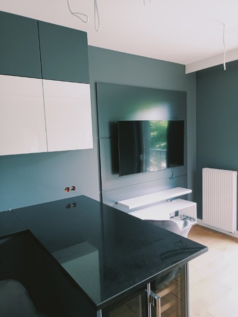 Nowoczesna aranżacja wnętrza z ciemnoszarą ścianą, białymi szafkami i czarnym blatem kuchennym, wiszący telewizor i biały grzejnik w tle.