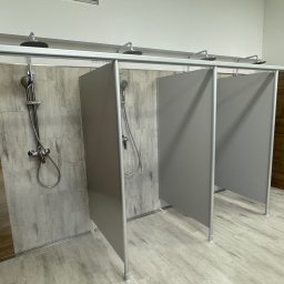 Gab-House Usługi Budowlane Damian Czarnocki - Nowoczesne kabiny prysznicowe z szarymi ściankami działowymi i drewnopodobnymi płytkami na ścianach i podłodze. Widoczne chromowane baterie i deszczownice.