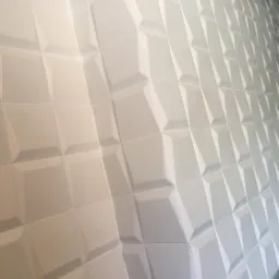 Biała ściana pokryta trójwymiarowymi panelami dekoracyjnymi o geometrycznym wzorze, ułożonymi pod kątem, z widocznym fragmentem szarej podłogi w dolnym rogu.