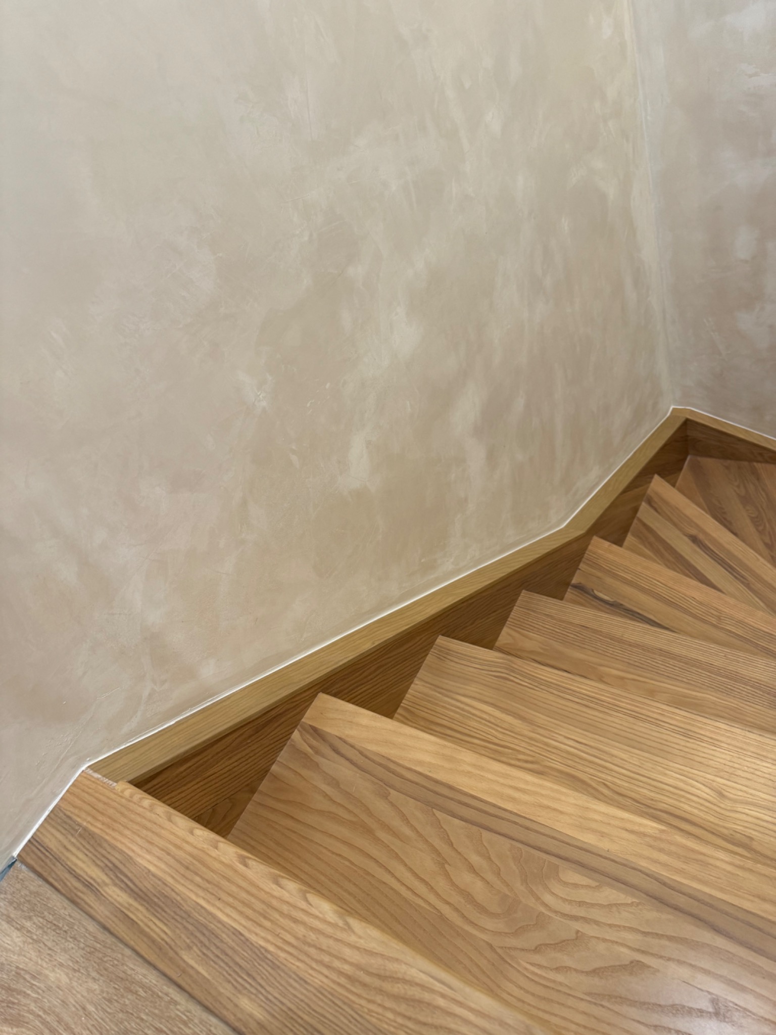 Drewniane schody spiralne z subtelnym oświetleniem i dekoracyjną ścianą w odcieniach beżu, tworzące eleganckie i nowoczesne wnętrze.