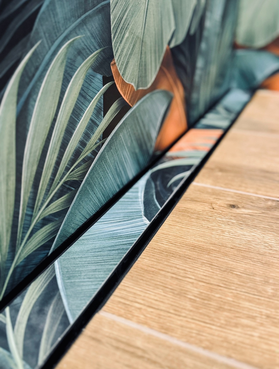 Detal wykończenia wnętrza: połączenie drewnopodobnych płytek podłogowych i ściany z motywem liści w odcieniach zieleni i pomarańczy, oddzielone czarną listwą.