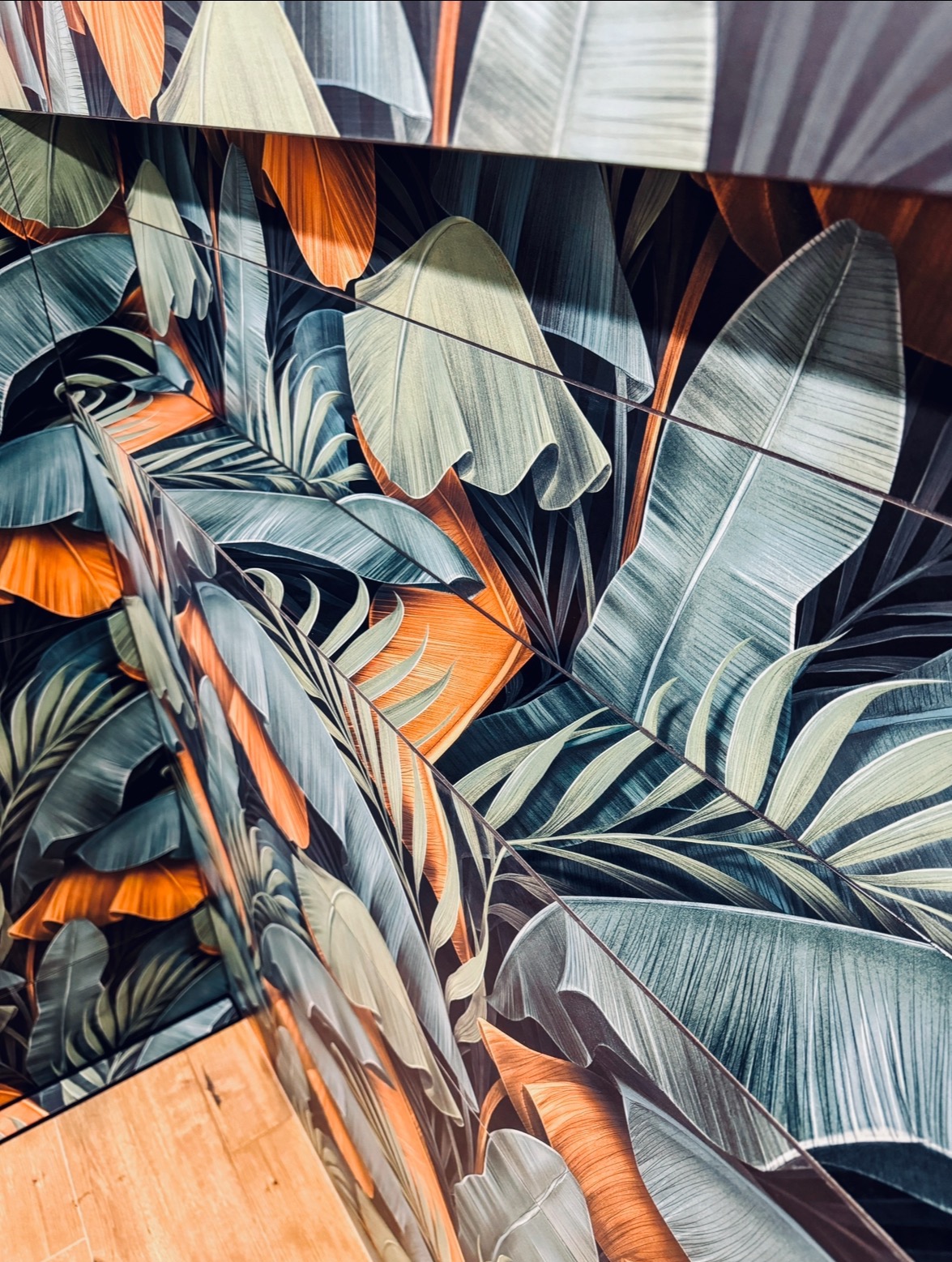 Ściana wykończona płytkami z motywem tropikalnych liści w odcieniach zieleni i pomarańczy, ułożonych pod kątem, z widoczną krawędzią podłogi.