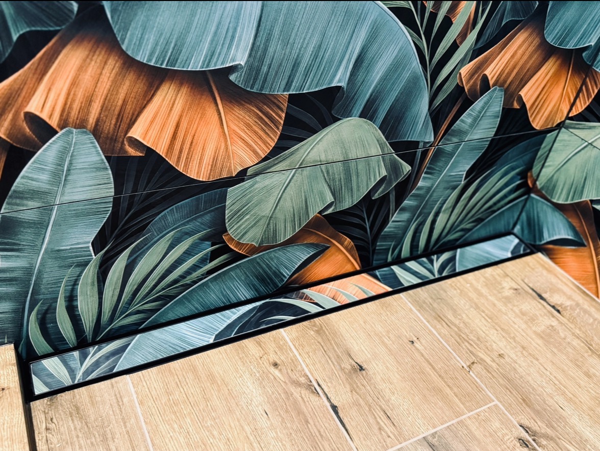 Wzorzysta ściana z motywem tropikalnych liści w odcieniach zieleni i pomarańczy, połączona z podłogą imitującą drewno, oddzielona czarną listwą.