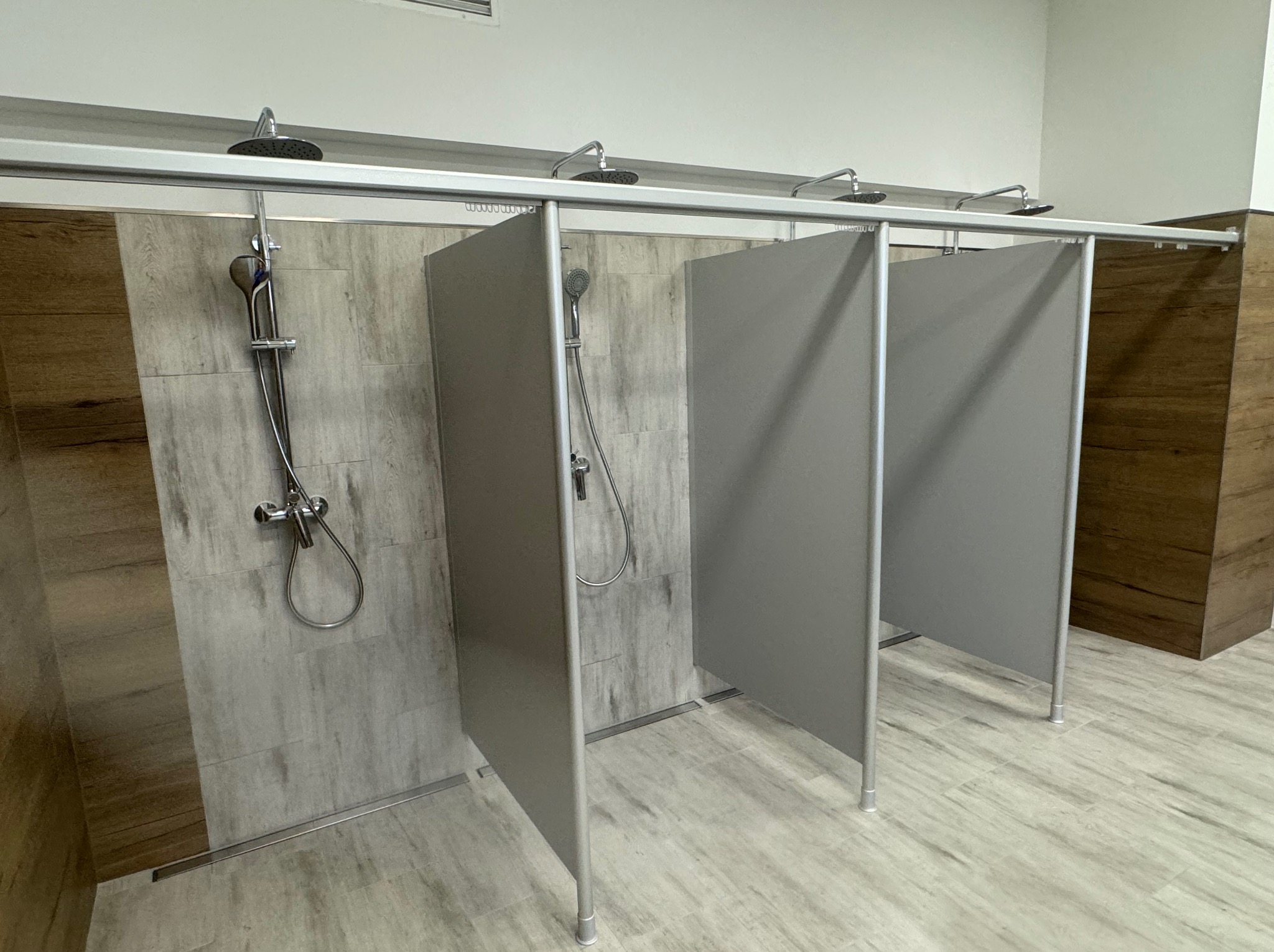 Nowoczesne kabiny prysznicowe z szarymi ściankami działowymi i drewnopodobnymi płytkami na ścianach i podłodze. Widoczne chromowane baterie i deszczownice.