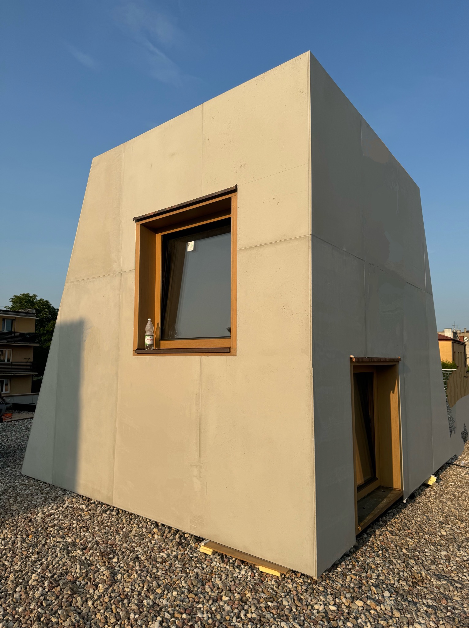 Nowoczesna, surowa bryła budynku z oknami w drewnianych ramach, ustawiona na żwirowym podłożu. Minimalistyczny design, jasne elewacje, błękitne niebo w tle.