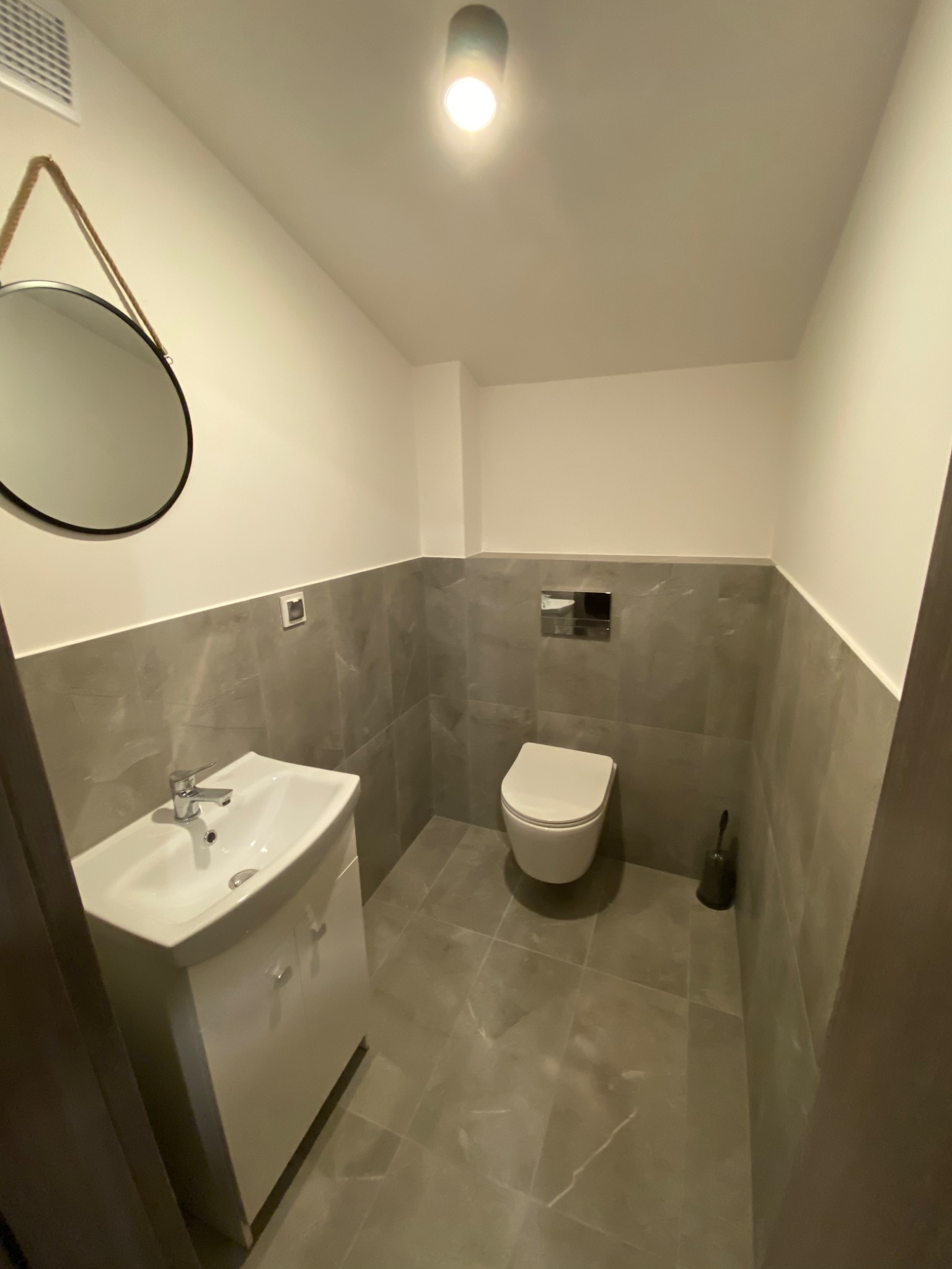 Nowoczesna łazienka wykończona w szaro-białej kolorystyce, z okrągłym lustrem na sznurze, białą umywalką z szafką, podwieszaną toaletą i szarymi płytkami imitującymi marmur.