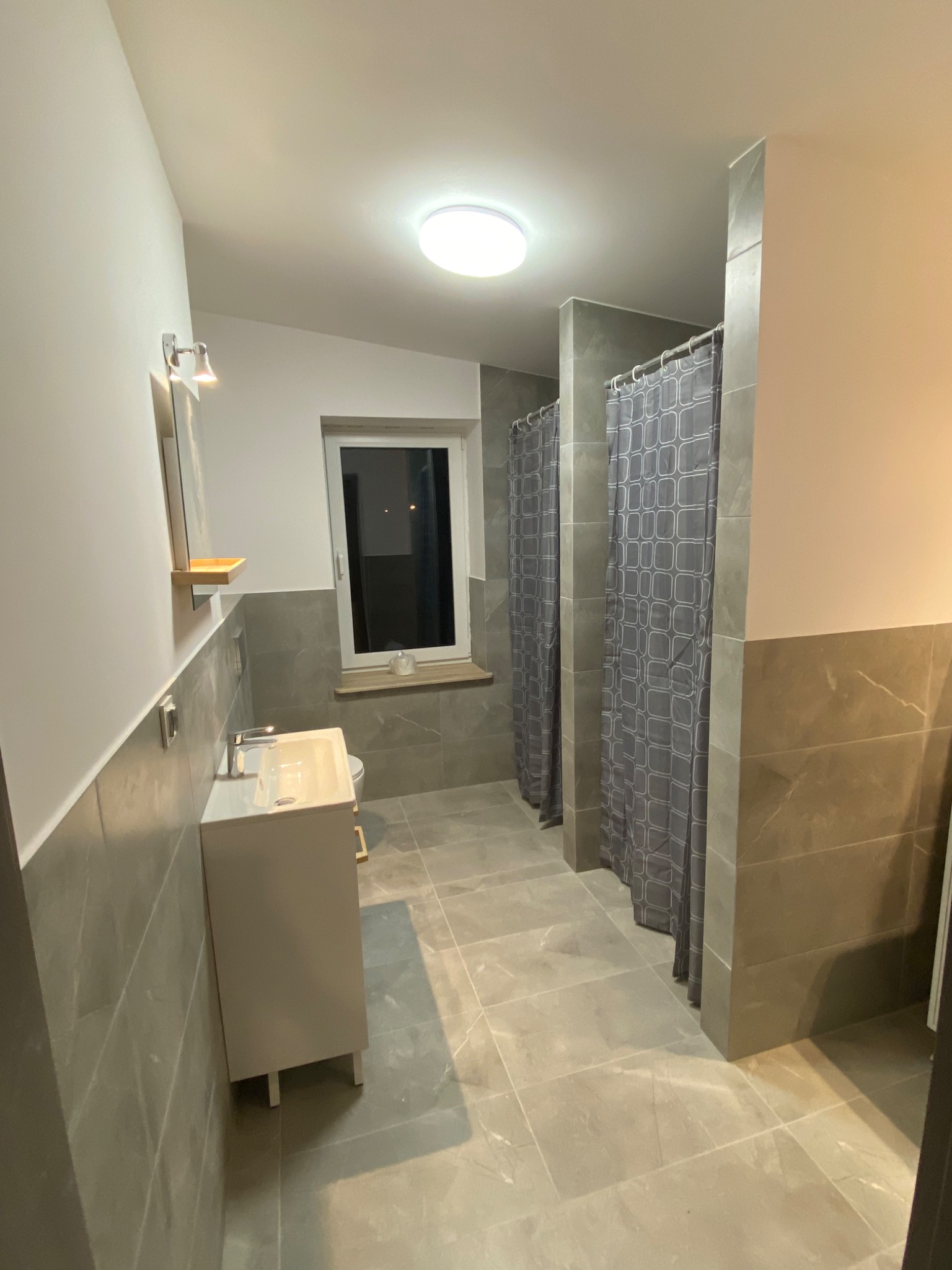 Wykończona łazienka w Mławie z szarymi płytkami imitującymi marmur na ścianach i podłodze, biała szafka pod umywalką i kabiny prysznicowe z szarymi zasłonami w geometryczny wzór.