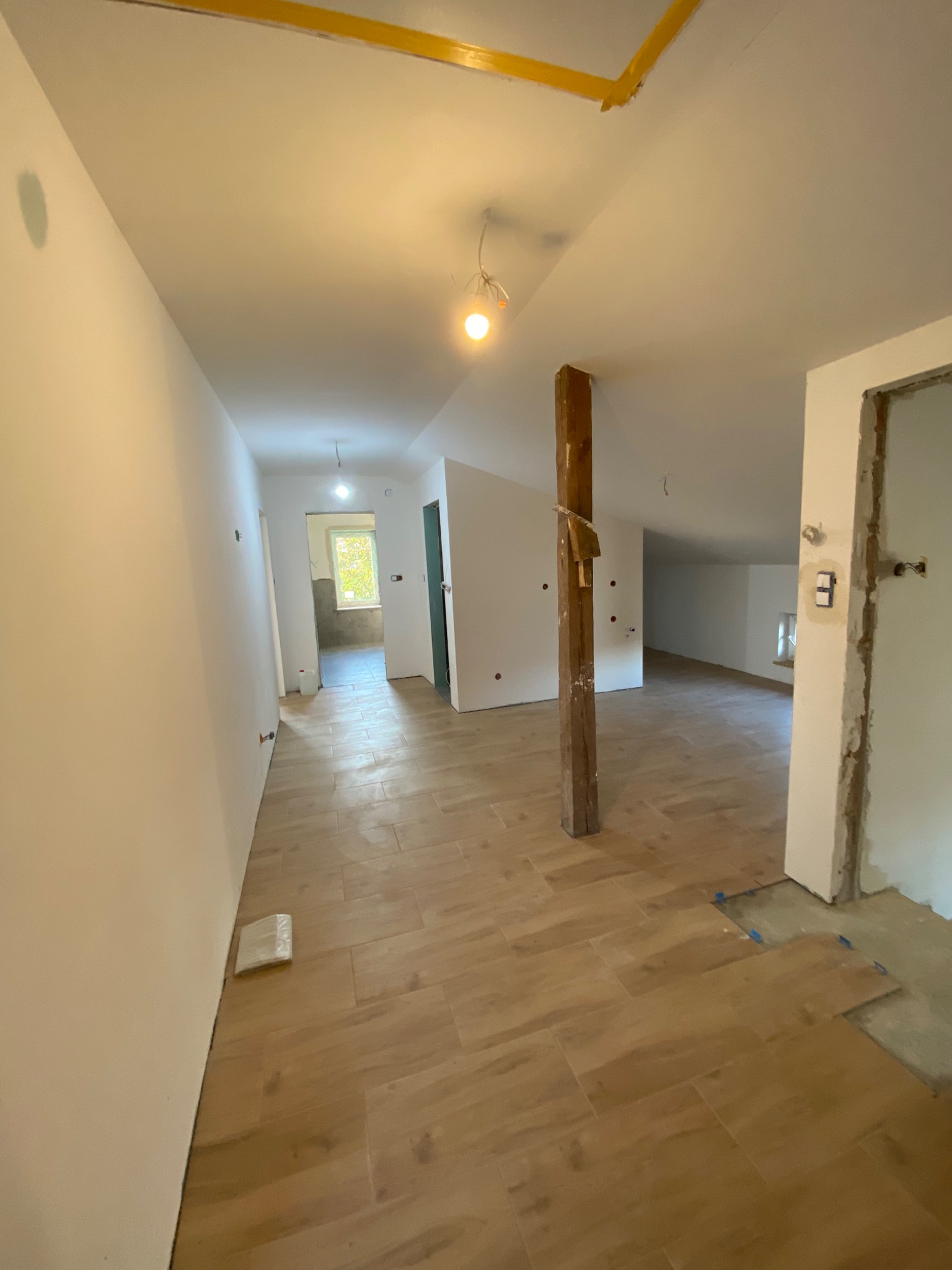 Wnętrze w trakcie remontu: świeżo pomalowane białe ściany, ułożona drewnopodobna podłoga, widoczny drewniany słup i fragmenty niezakończonych prac wykończeniowych przy drzwiach.