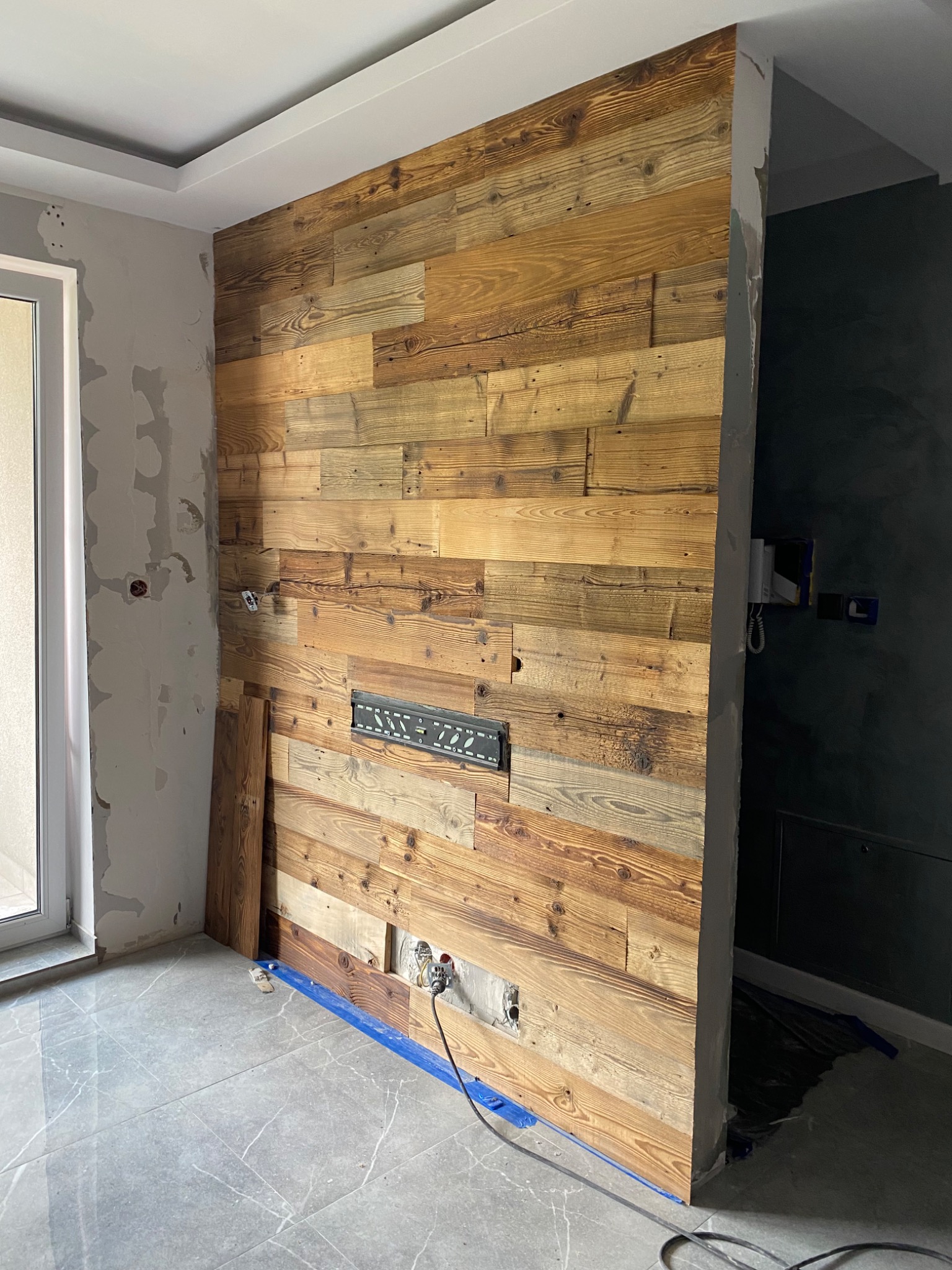 Ściana wykończona drewnem z recyklingu w trakcie montażu, widoczne puszki elektryczne, nieotynkowane ściany i zabezpieczona podłoga, element dekoracyjny wnętrza w surowym stylu.