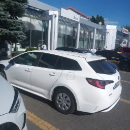 Biała Toyota Corolla kombi na parkingu przed salonem samochodowym Żerań w słoneczny dzień, widoczne inne samochody i logo Toyota.