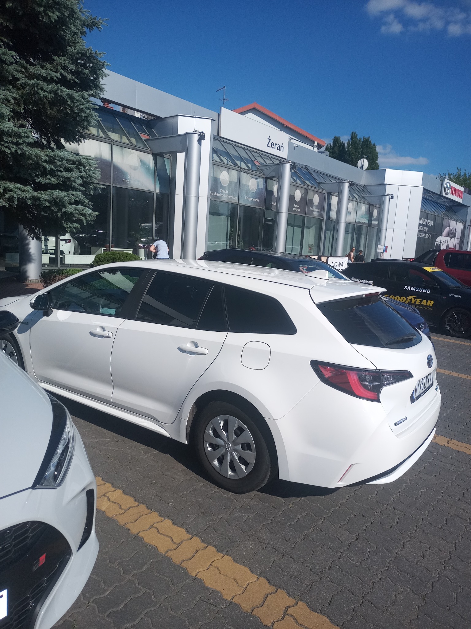 Biała Toyota Corolla kombi na parkingu przed salonem samochodowym Żerań w słoneczny dzień, widoczne inne samochody i logo Toyota.