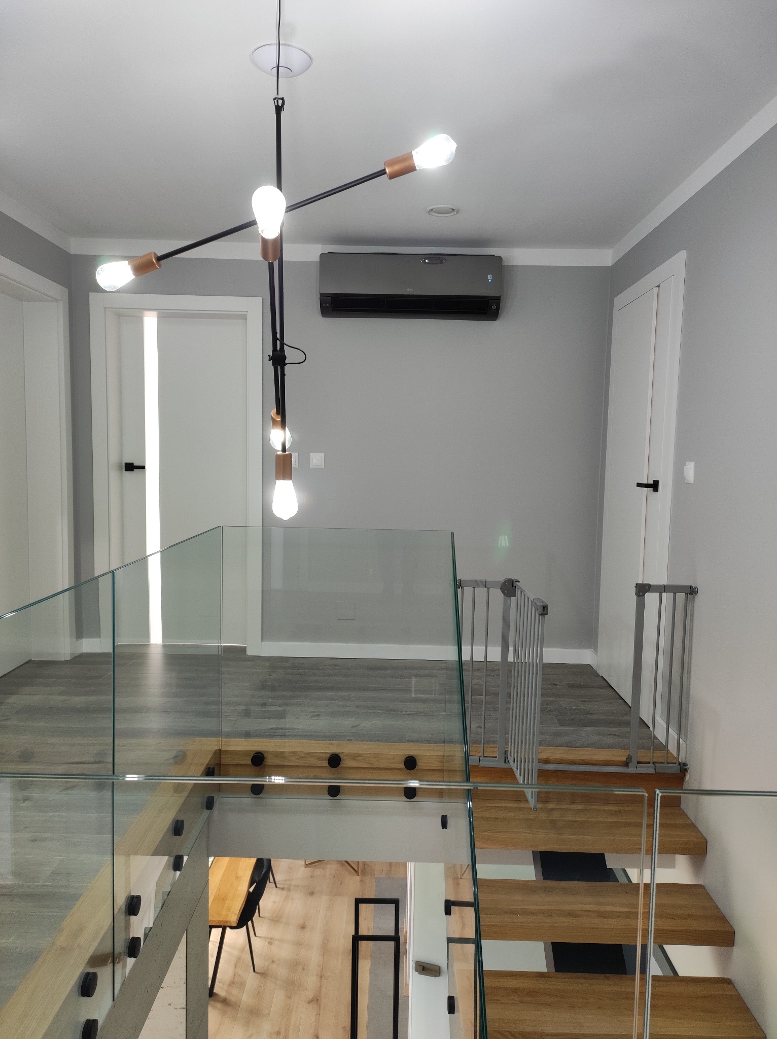 Wnętrze domu z zamontowaną, szarą klimatyzacją ścienną nad schodami z drewnianymi stopniami i szklaną balustradą, widoczne zabezpieczenia schodów dla dzieci.