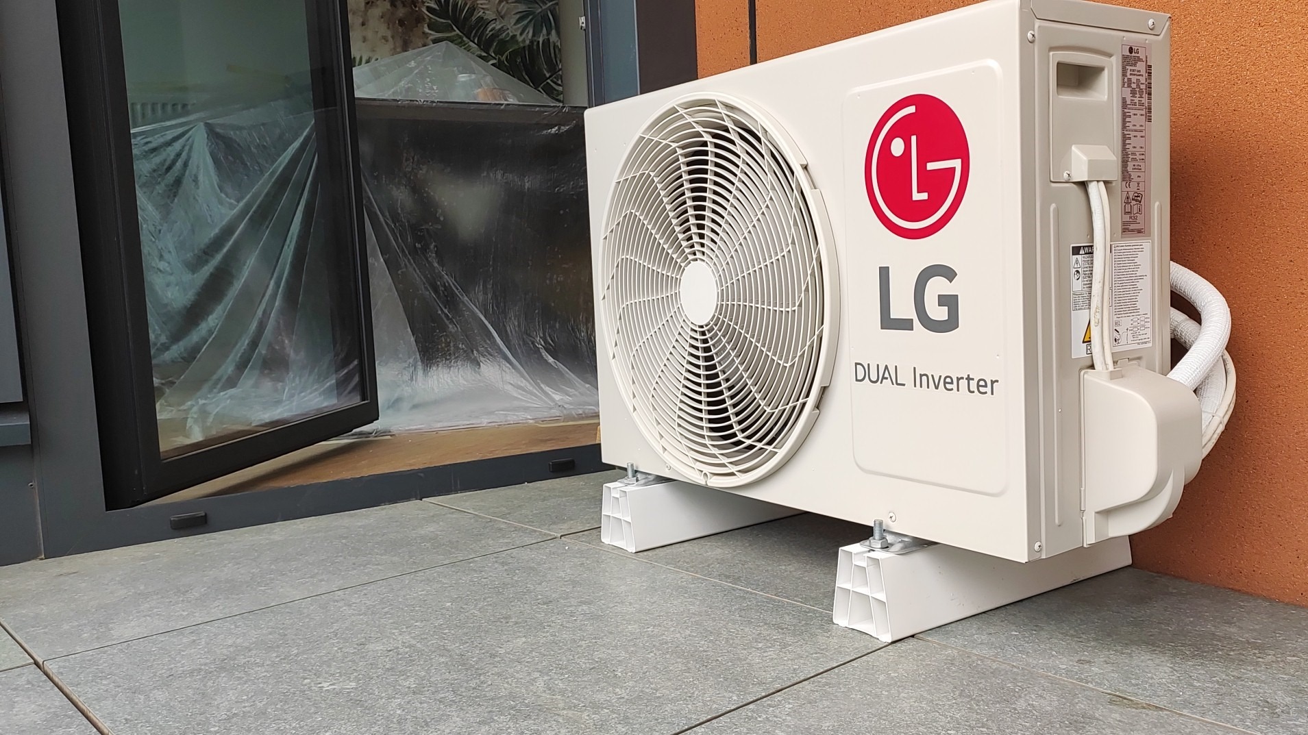 Zewnętrzna jednostka klimatyzacji LG Dual Inverter, zamontowana na białych wspornikach na szarym tarasie, z otwartym oknem w tle zabezpieczonym folią.