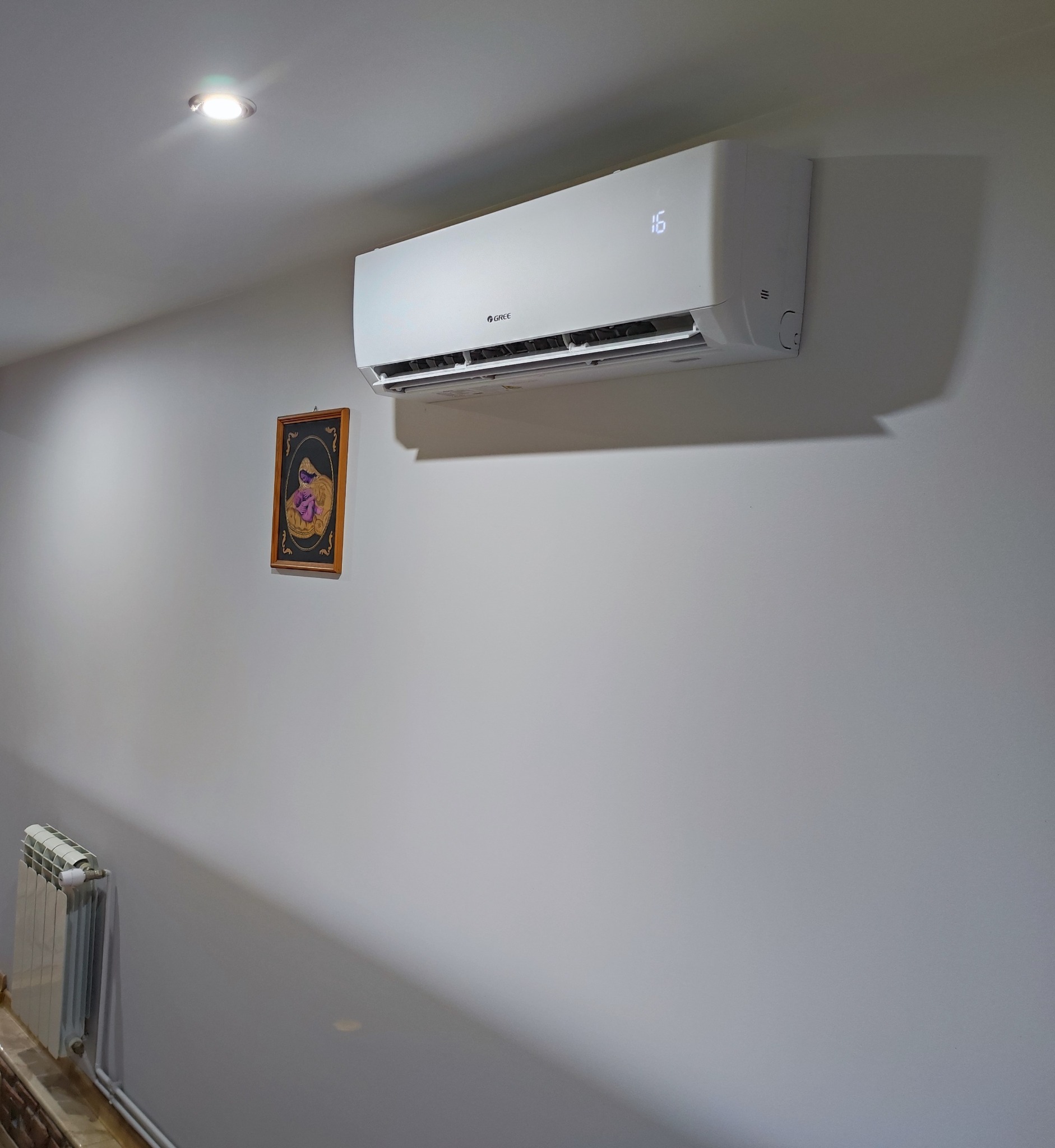 Biały klimatyzator marki Gree zamontowany na ścianie w pomieszczeniu, obok wiszący obrazek w drewnianej ramie i grzejnik.