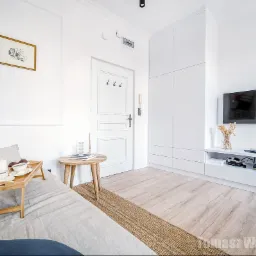 Jasne wnętrze pokoju dziennego z sofą, stolikiem kawowym z przekąskami, telewizorem i białymi meblami na tle białych ścian z dekoracyjną listwą.