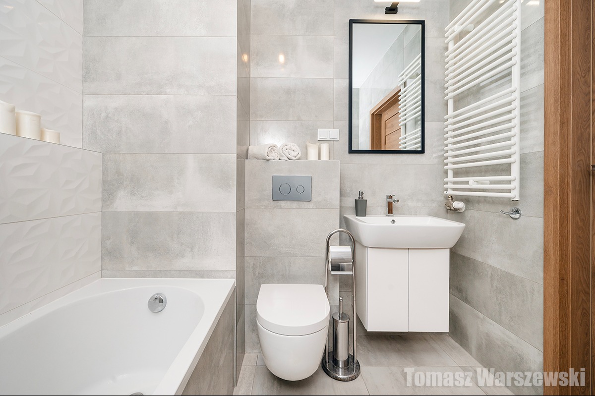 Nowoczesna łazienka w odcieniach szarości z białą armaturą, grzejnikiem drabinkowym i lustrem w czarnej ramie, oświetlona lampą nad lustrem.