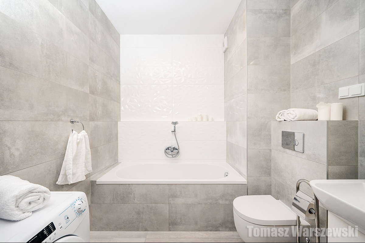 Nowoczesna łazienka w odcieniach szarości i bieli, z wanną, toaletą, umywalką i pralką. Na ścianach duże, prostokątne płytki imitujące beton. Nad wanną białe płytki z delikatnym wzorem 3D. Widoczny...