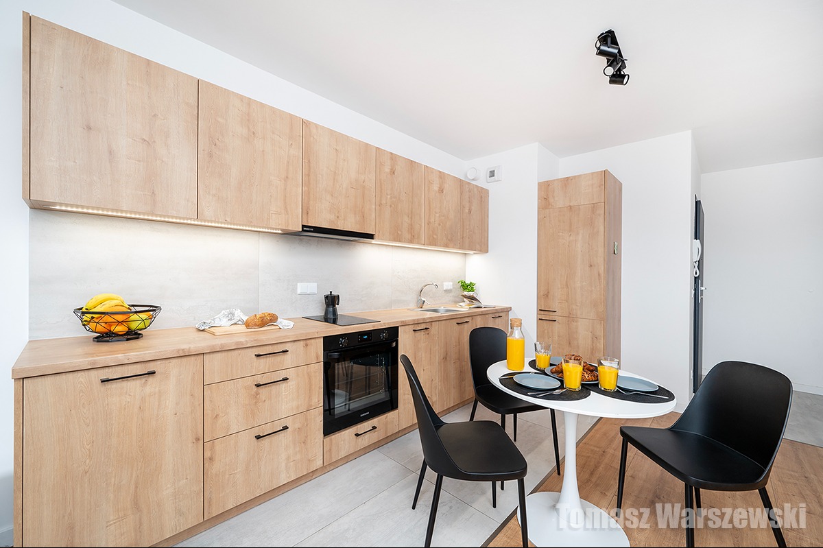 Jasna kuchnia z drewnianymi szafkami, czarnymi uchwytami i okrągłym stołem z czarnymi krzesłami, na blacie koszyk z owocami i deska z pieczywem.