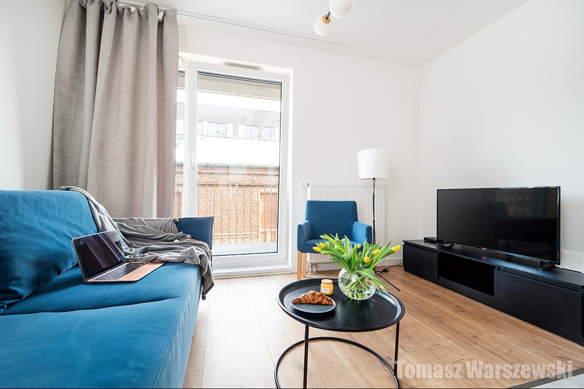 Jasne wnętrze pokoju dziennego z błękitną sofą, fotelem i czarnym stolikiem kawowym z croissantem i tulipanami, laptop na sofie, widok na ceglany budynek za oknem.
