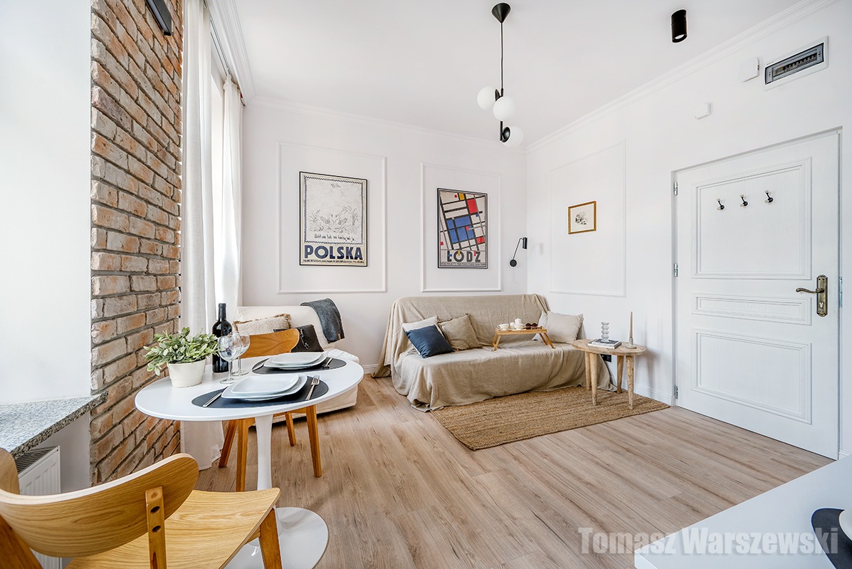 Jasne wnętrze mieszkania z ceglaną ścianą, okrągłym stołem z nakryciem, sofą z poduszkami i plakatami na ścianach. Widoczny fragment drewnianego krzesła i dywanik z juty.
