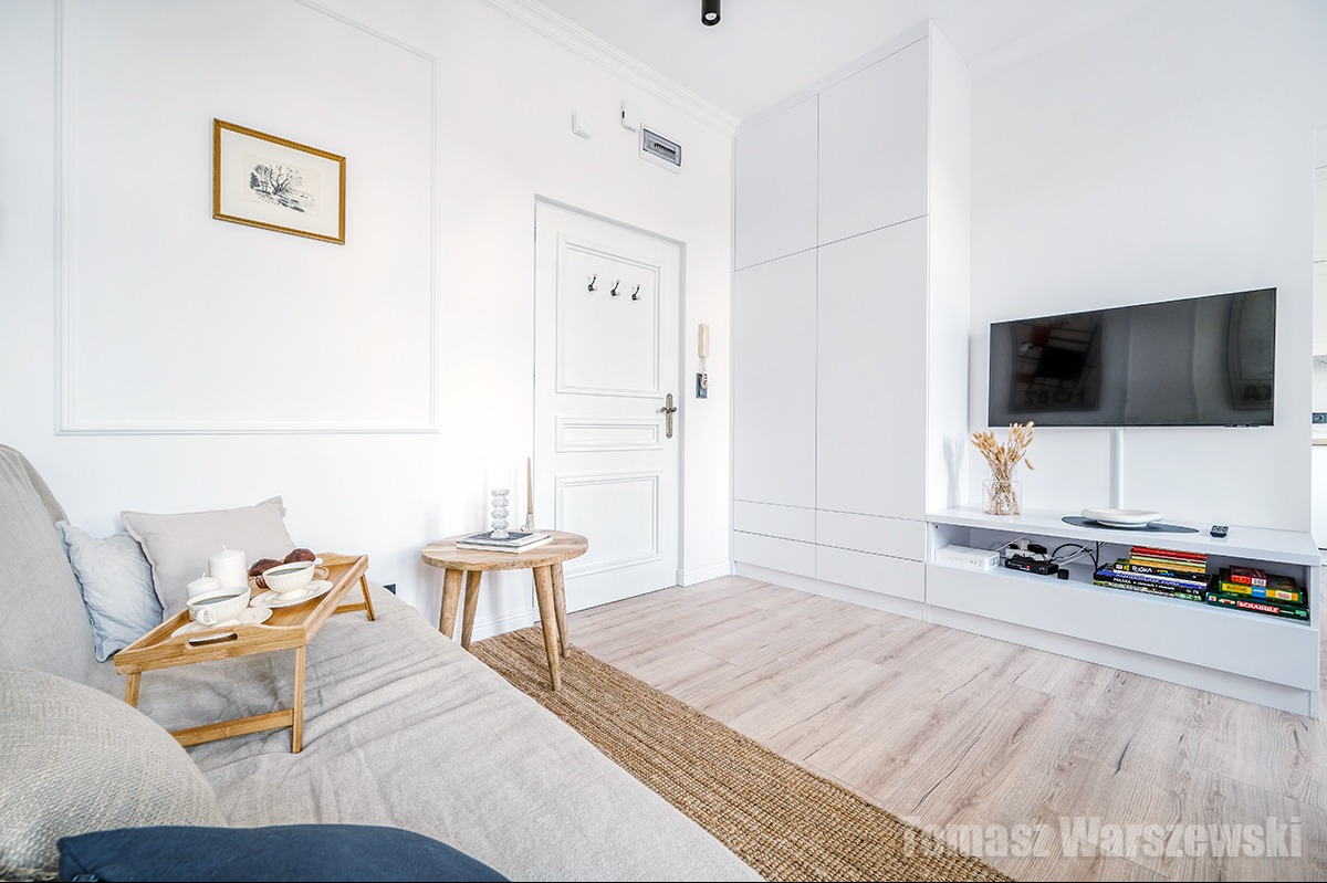 Jasne wnętrze pokoju dziennego z sofą, stolikiem kawowym z przekąskami, telewizorem i białymi meblami na tle białych ścian z dekoracyjną listwą.