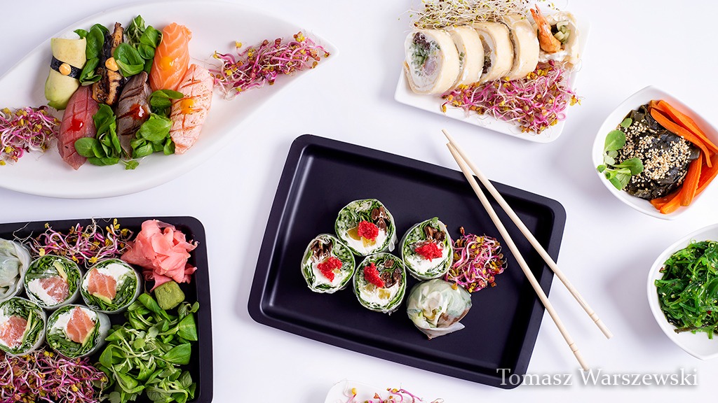 Kompozycja różnorodnych dań kuchni azjatyckiej, w tym sushi i sajgonki, podanych na talerzach i tacach na białym tle, z dekoracją z kiełków.