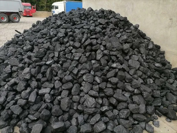 Góra czarnego węgla kamiennego na placu składowym, z widocznymi w tle samochodami ciężarowymi.