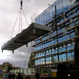 Montaż prefabrykowanego elementu elewacji na budynku biurowym z przeszkloną fasadą, widoczne rusztowania i pracownicy.