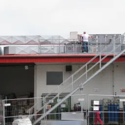 Instalacja wentylacyjna przemysłowa na dachu budynku, pracownicy na metalowych schodach przeprowadzają kontrolę, widoczne przewody wentylacyjne i urządzenia klimatyzacyjne.