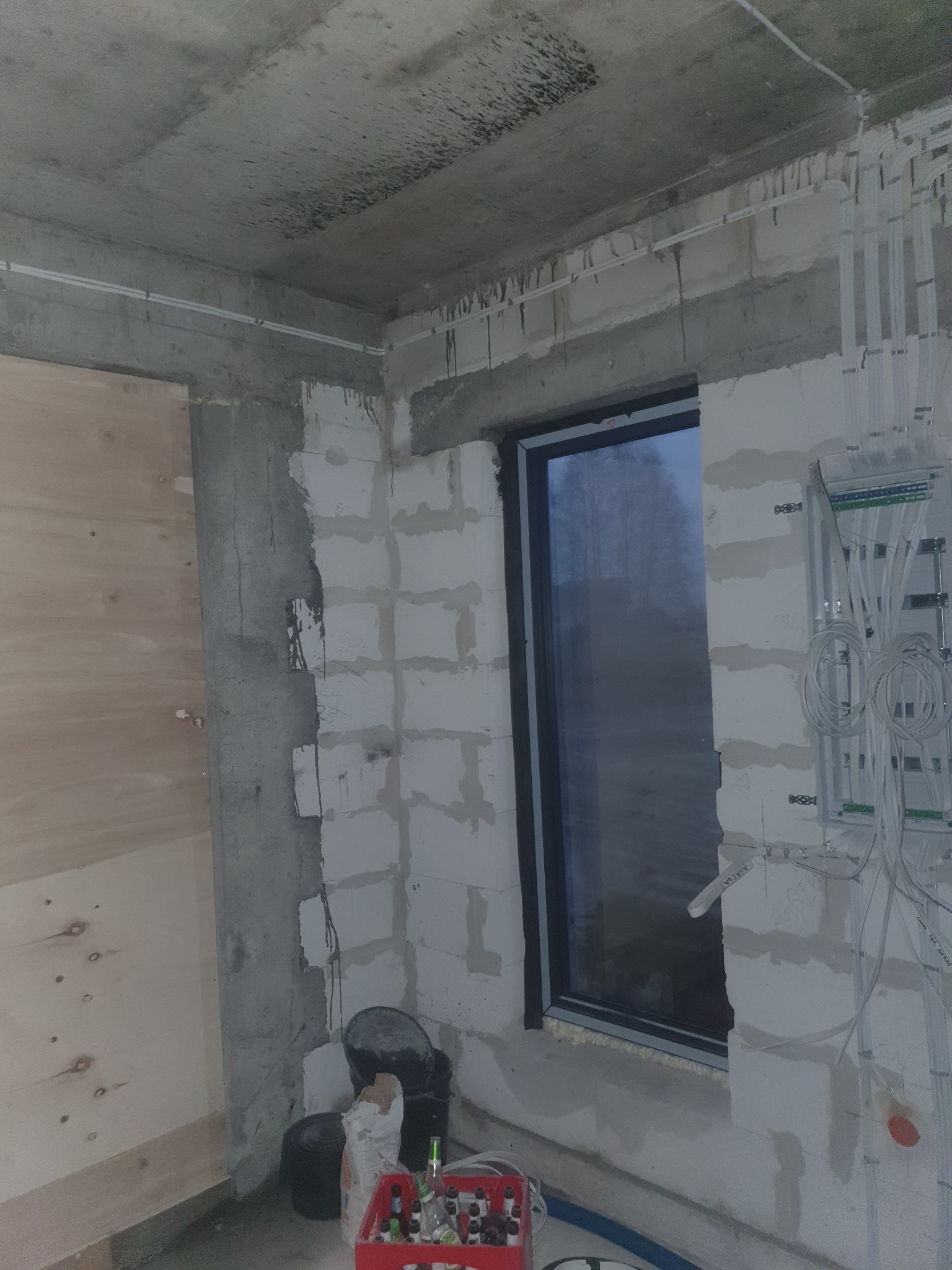 Surowy stan wnętrza w trakcie prac wykończeniowych, widoczne kable elektryczne prowadzone po ścianach, skrzynka rozdzielcza, okno z czarną ramą oraz materiały budowlane i puste butelki po napojach...