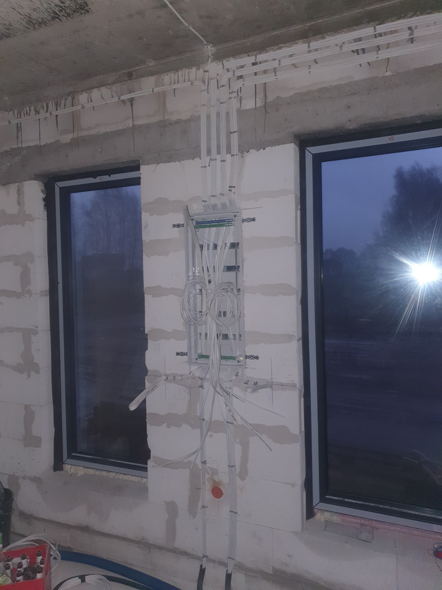 Rozdzielnica elektryczna z podłączonymi przewodami na ścianie z betonu komórkowego, widoczne okna z ciemnymi szybami, instalacja przewodów w rurkach PCV.