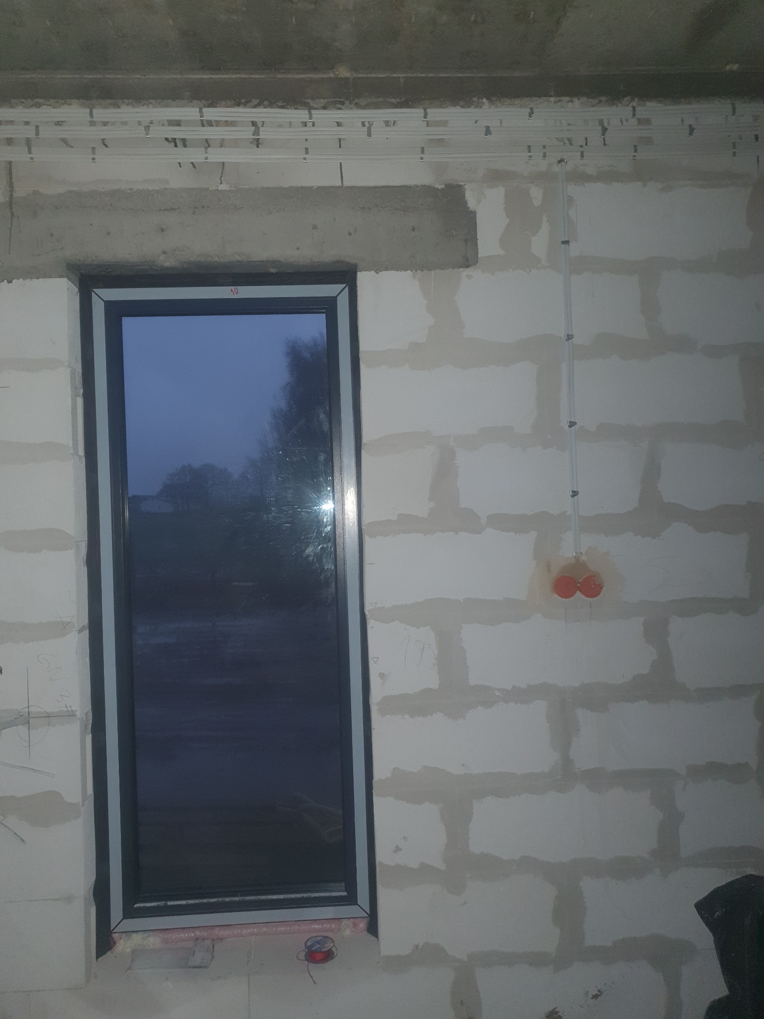 Surowy stan wnętrza z widocznym oknem, instalacją elektryczną w peszlu i puszkami pod gniazdka na ścianie z bloczków, listwy montażowe pod sufitem.
