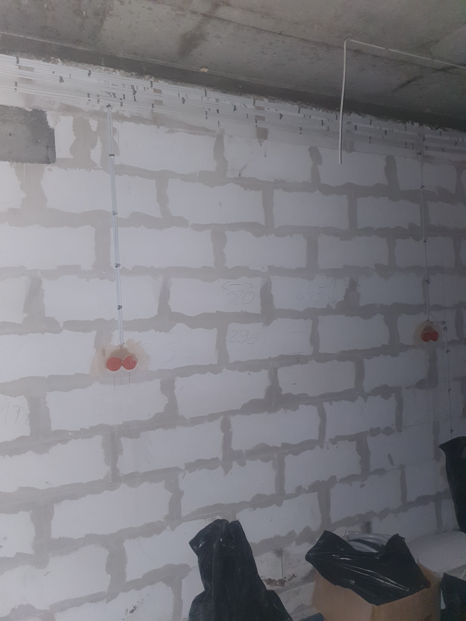 Ściana z bloków betonowych w trakcie prac instalacyjnych, widoczne koryta kablowe pod sufitem, pionowe rury instalacyjne z puszkami elektrycznymi, oraz napisy ołówkiem na ścianie.