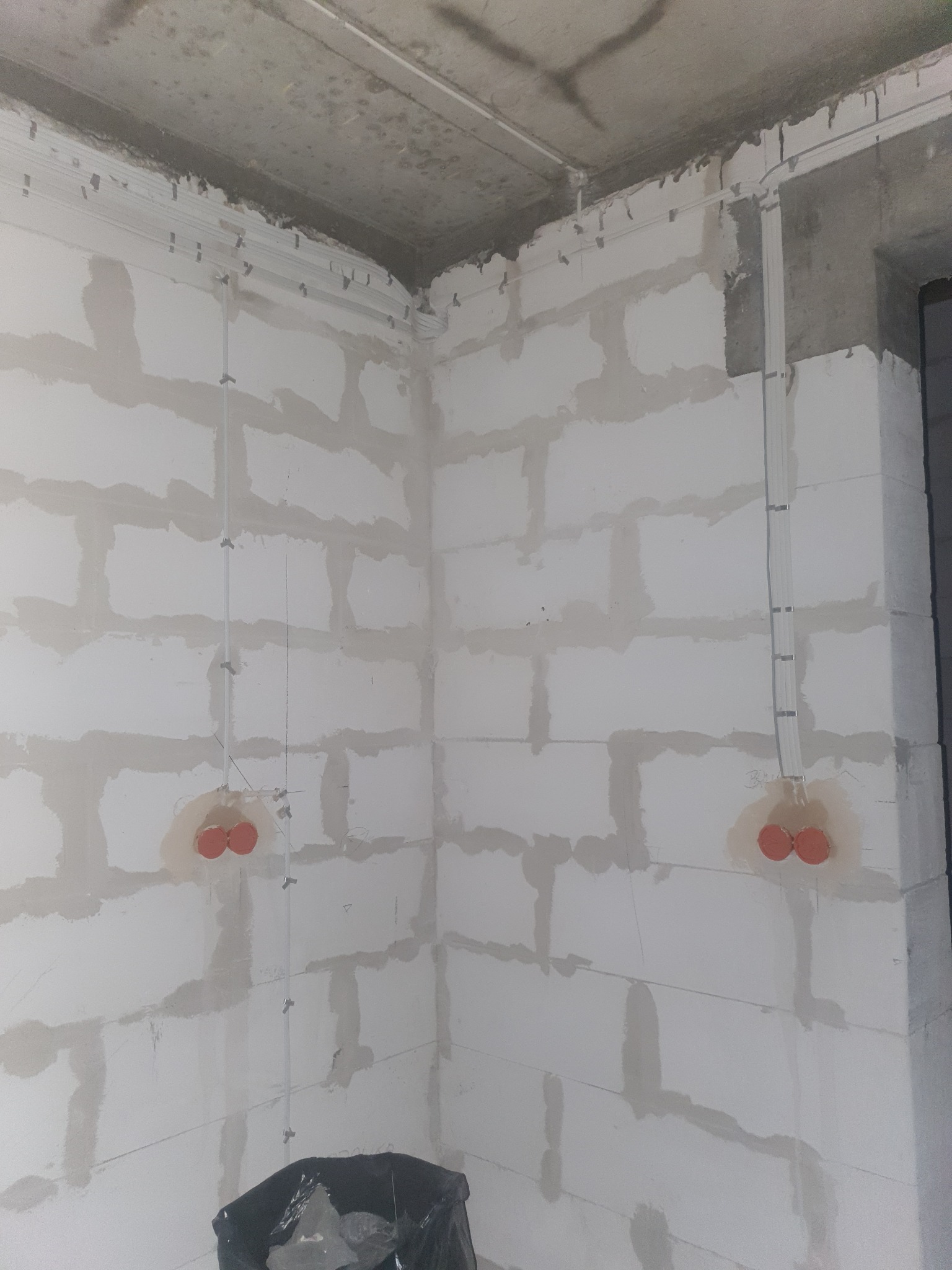 Surowy stan ściany z betonu komórkowego w trakcie prac elektrycznych: widoczne kable w peszlach i puszki instalacyjne z pomarańczowymi kołnierzami, mocowane do ściany za pomocą uchwytów.