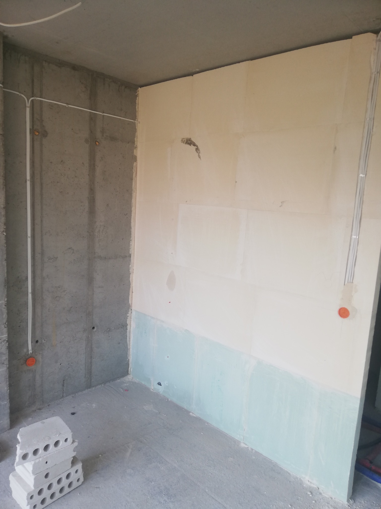 Surowy betonowy narożnik pokoju z poprowadzonymi przewodami elektrycznymi w białych rurkach, ściany częściowo wykończone płytami gipsowymi i pomalowane na biało i błękitno, na podłodze leżą bloczki...