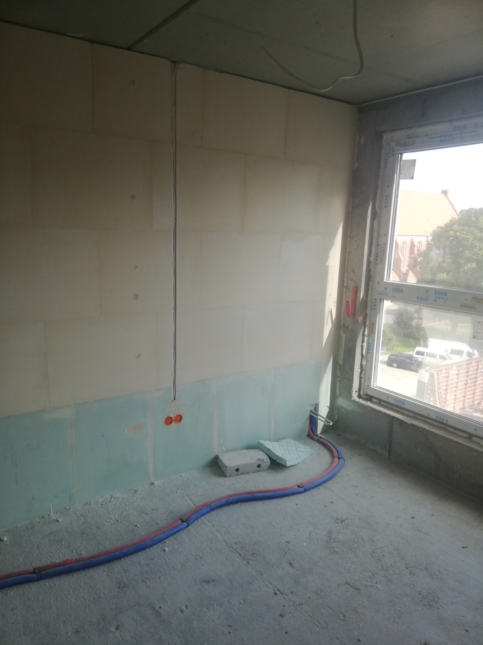 Surowy stan wnętrza w trakcie prac wykończeniowych; widoczne przewody elektryczne prowadzone po ścianie, puszki pod gniazdka oraz rury grzewcze ułożone na podłodze przy oknie.