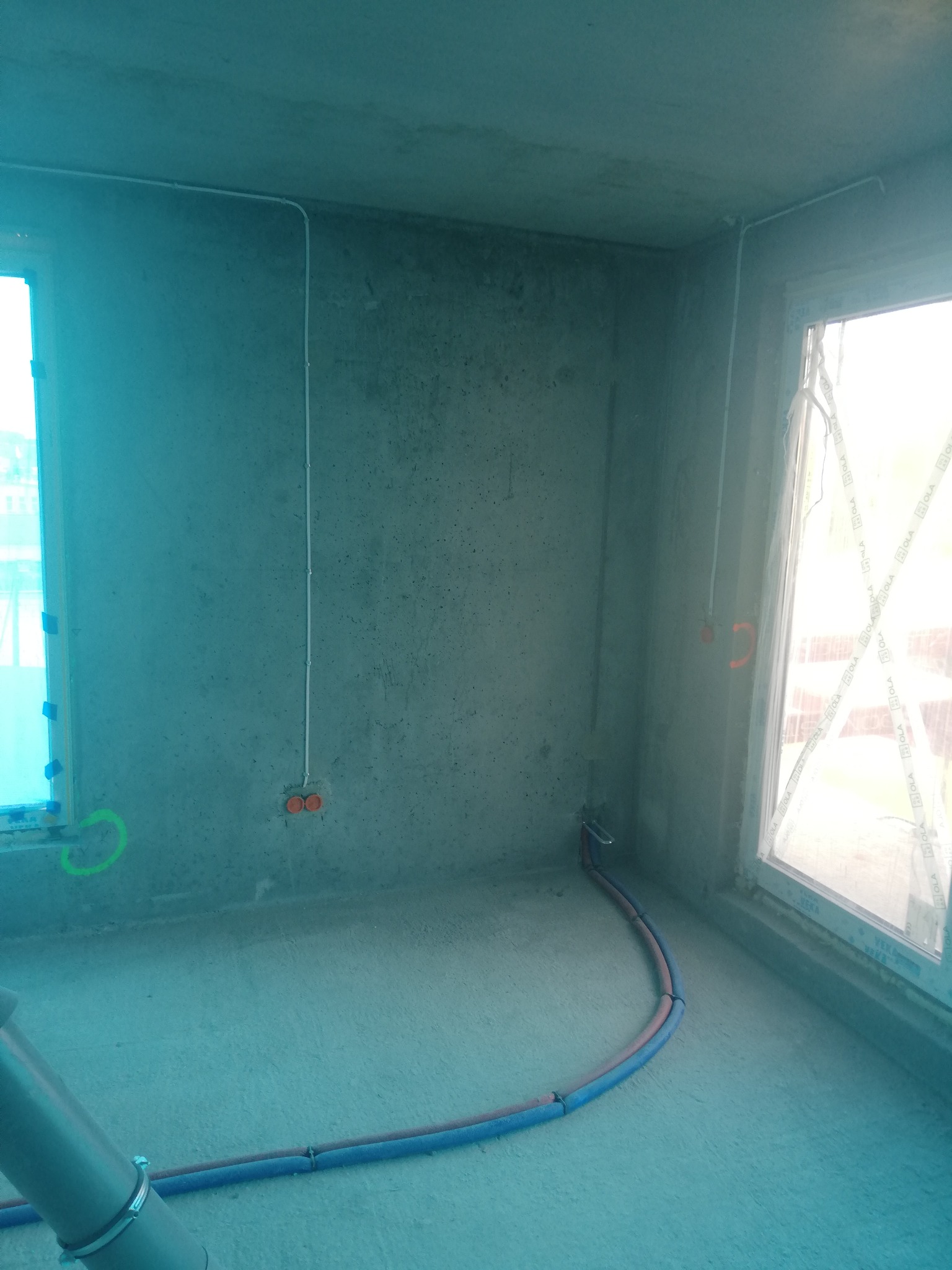 Surowy stan wykończeniowy pokoju z widocznymi przewodami elektrycznymi w białych listwach na ścianach, puszkami instalacyjnymi i rurami grzewczymi przy podłodze, okna zabezpieczone folią.