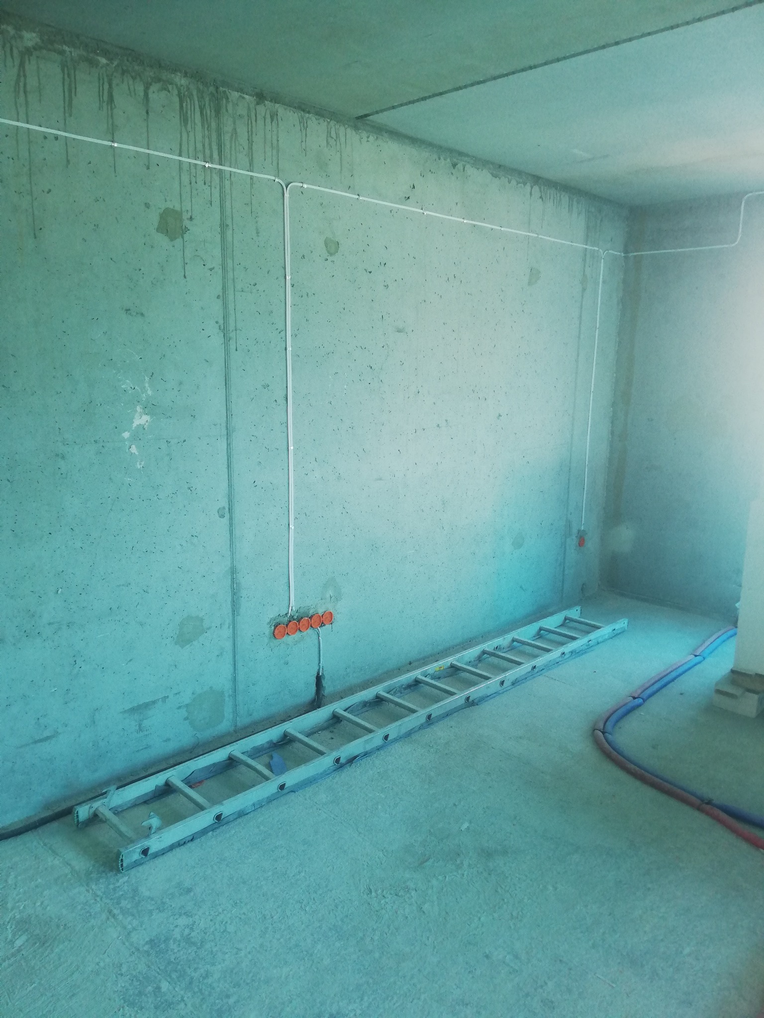 Surowy pokój z betonowymi ścianami, widoczne białe kable elektryczne prowadzone po ścianie i pomarańczowe puszki instalacyjne, na podłodze rozłożona aluminiowa drabina i rury w izolacji.