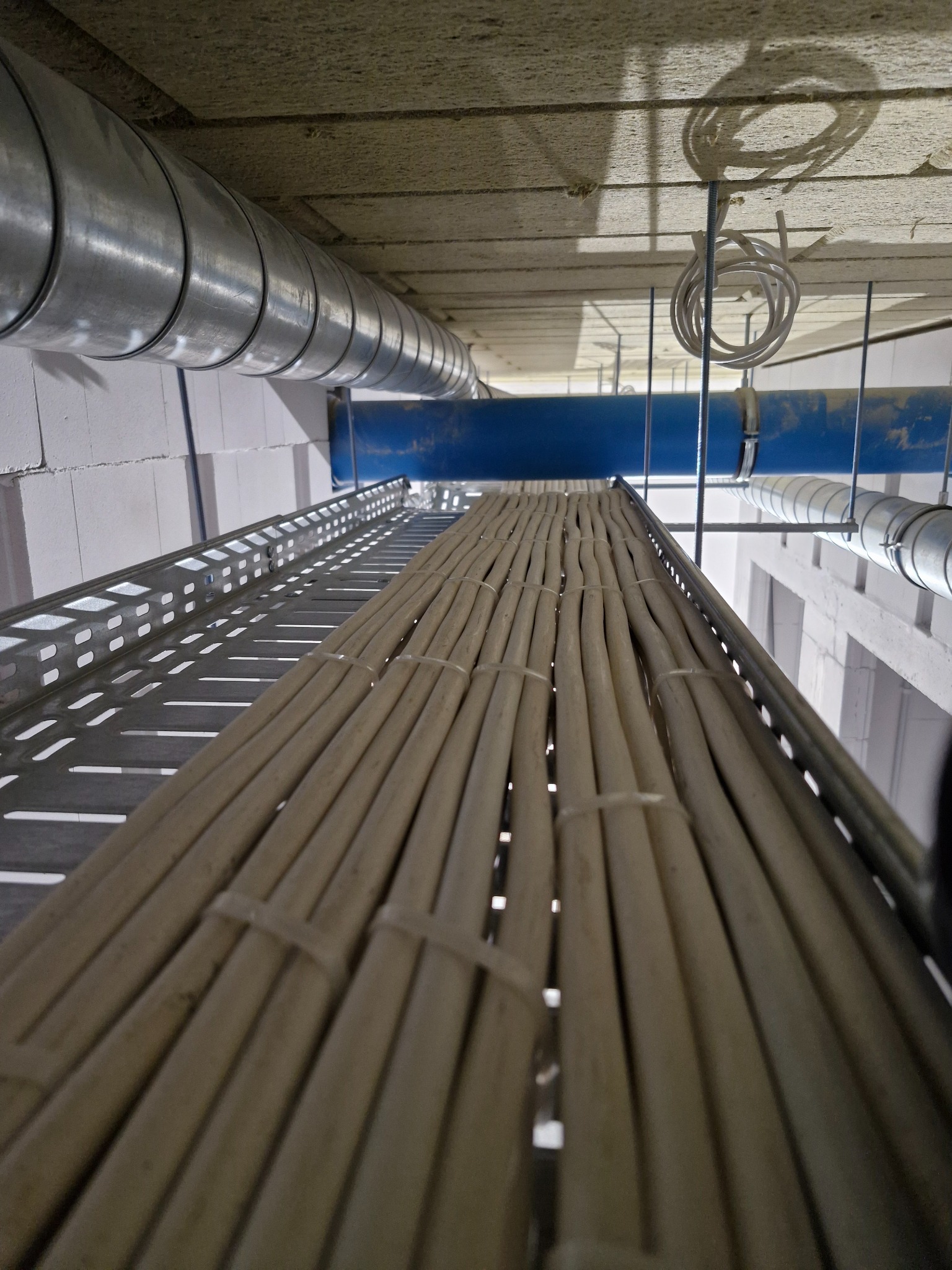 Kable elektryczne ułożone równolegle na perforowanej metalowej korycie kablowej pod sufitem, zabezpieczone opaskami zaciskowymi, widoczne rury wentylacyjne i instalacyjne.