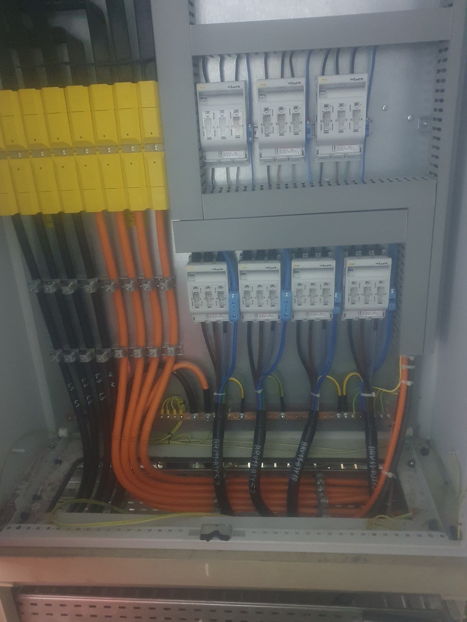 Wnętrze rozdzielnicy elektrycznej z ułożonymi przewodami o różnych kolorach (pomarańczowe, czarne, niebieskie, żółte) i zabezpieczeniami marki Wlux.